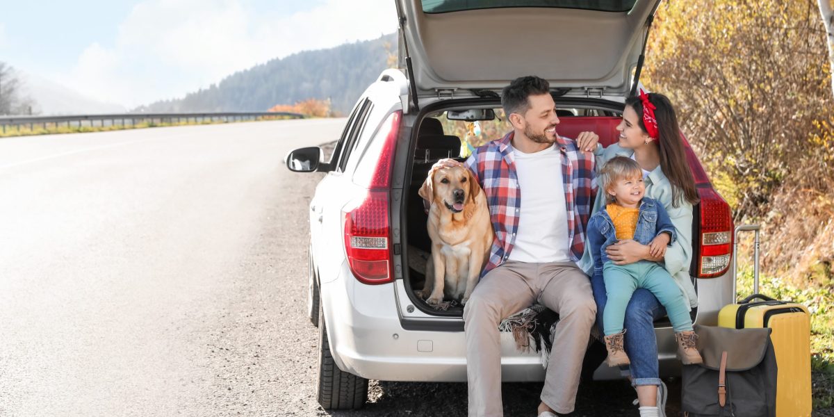 Parents, their daughter and dog sitting in car trunk near road, space for text. Family traveling with pet